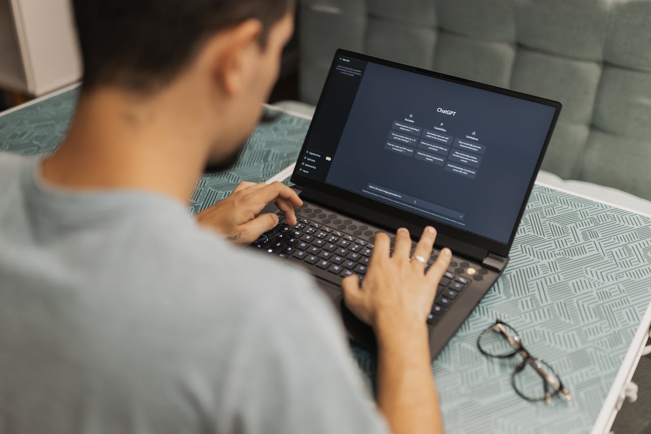 heros-img Man using laptop with AI interface, typing with focused attention. Indoors with eyewear beside.