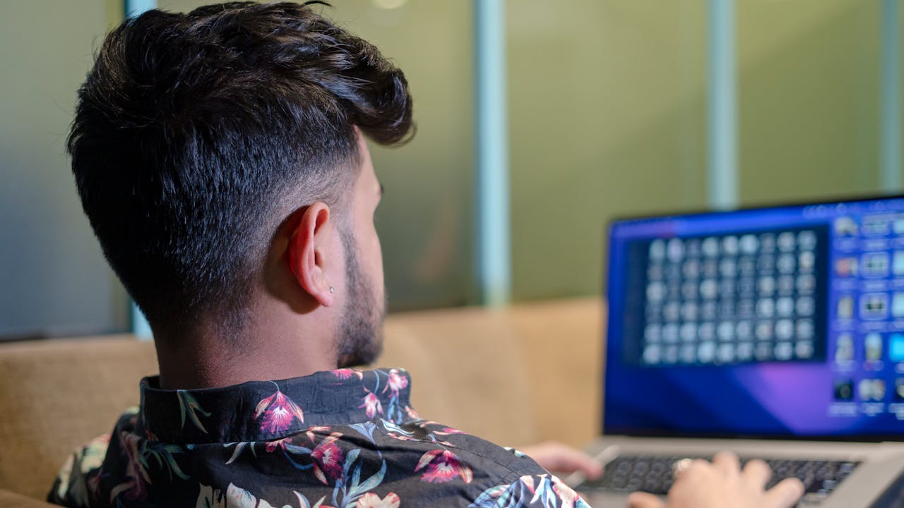 about-us-02 Man with short hair using a laptop indoors, viewed from behind. Floral shirt visible.